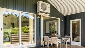 A modern outdoor patio with a wooden deck, a dining table with chairs, and a large sliding glass door leading to a garden.