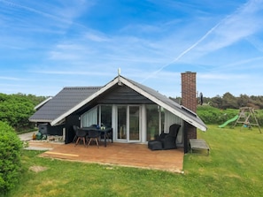 Ein Haus mit geneigtem Dach, einer Terrasse mit Gartenmöbeln, einem Schornstein und einem Spielbereich im Hintergrund.