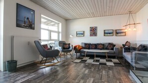 Un salon spacieux, doté d’un grand canapé, d’un fauteuil à bascule et d’une table basse. Plusieurs œuvres d’art encadrées ornent les murs, et une fenêtre laisse entrer la lumière naturelle.