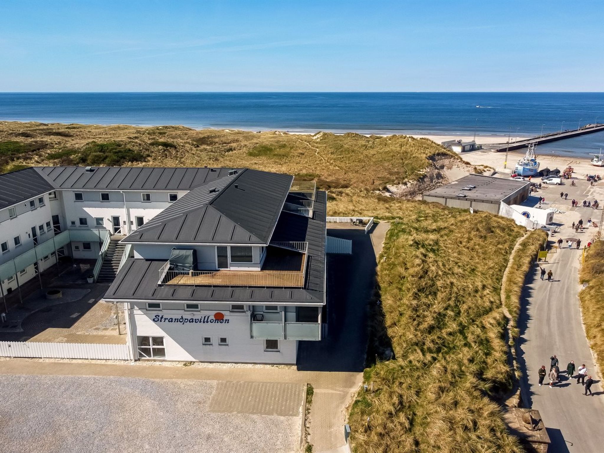 Un bâtiment moderne doté d’un toit plat et de plusieurs étages, situé près d’une plage avec une jetée et des bateaux.