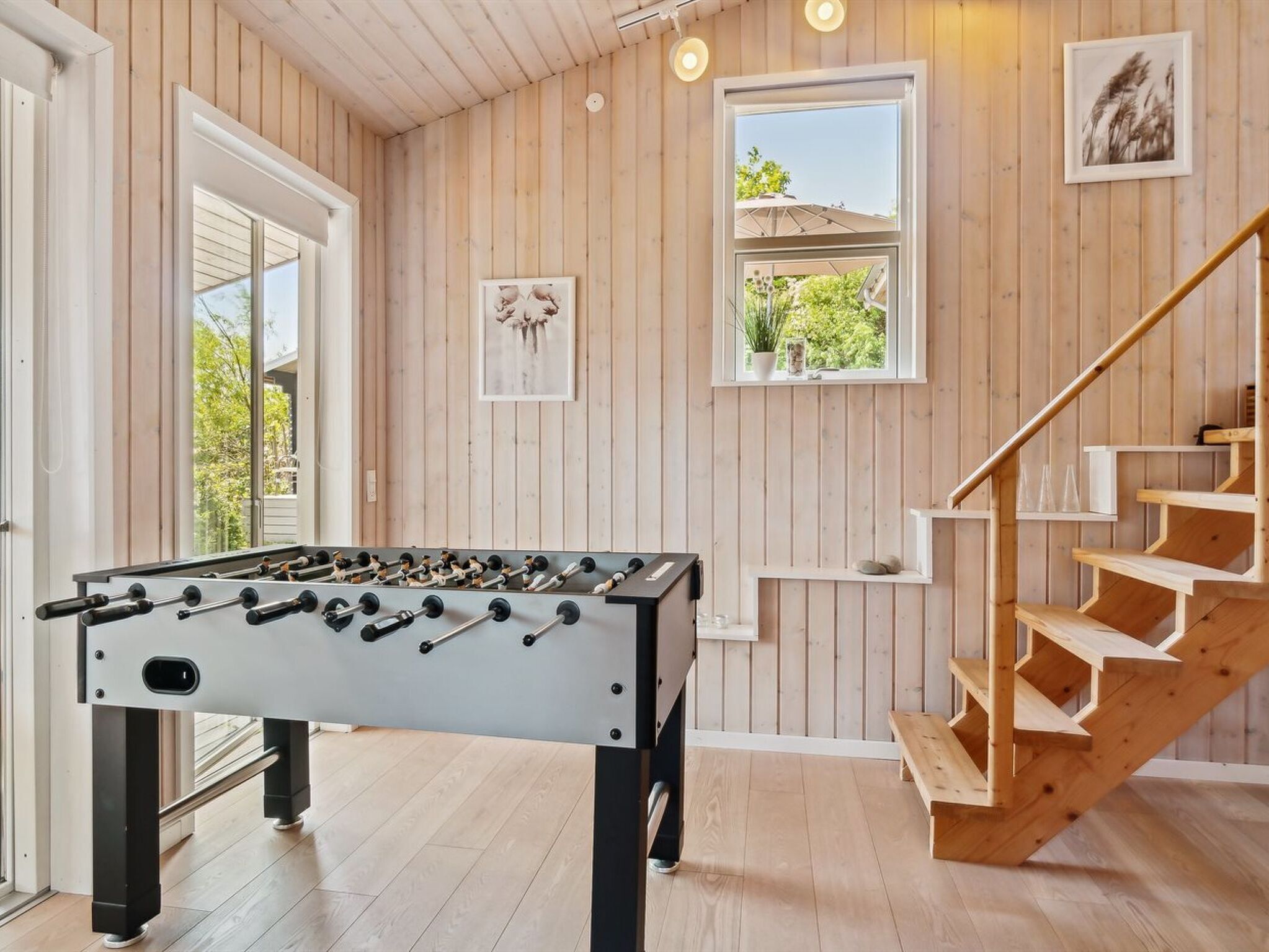 A room with a foosball table, wooden staircase, and a window with a view of greenery.