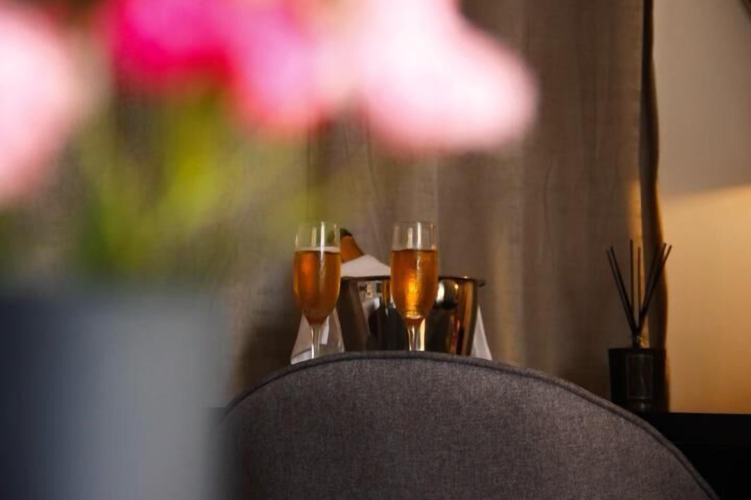 A table with two glasses of amber liquid, possibly wine, and a vase with a pink flower.