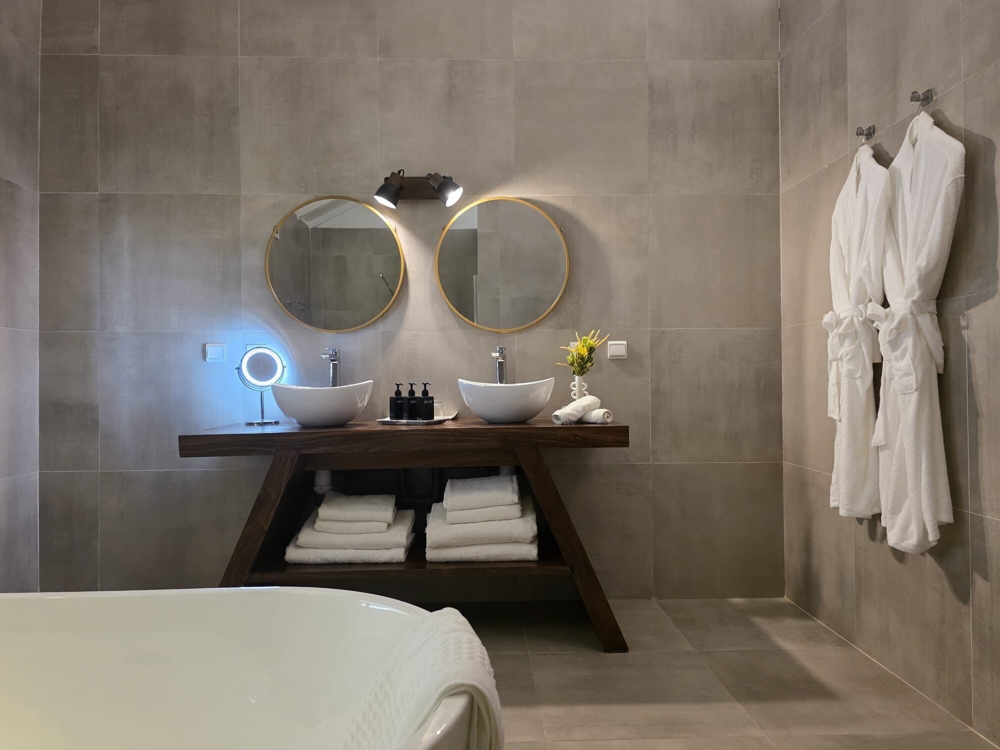 Un baño moderno con dos espejos redondos, un mueble de tocador de madera, un lavabo sobre encimera y un perchero con batas.