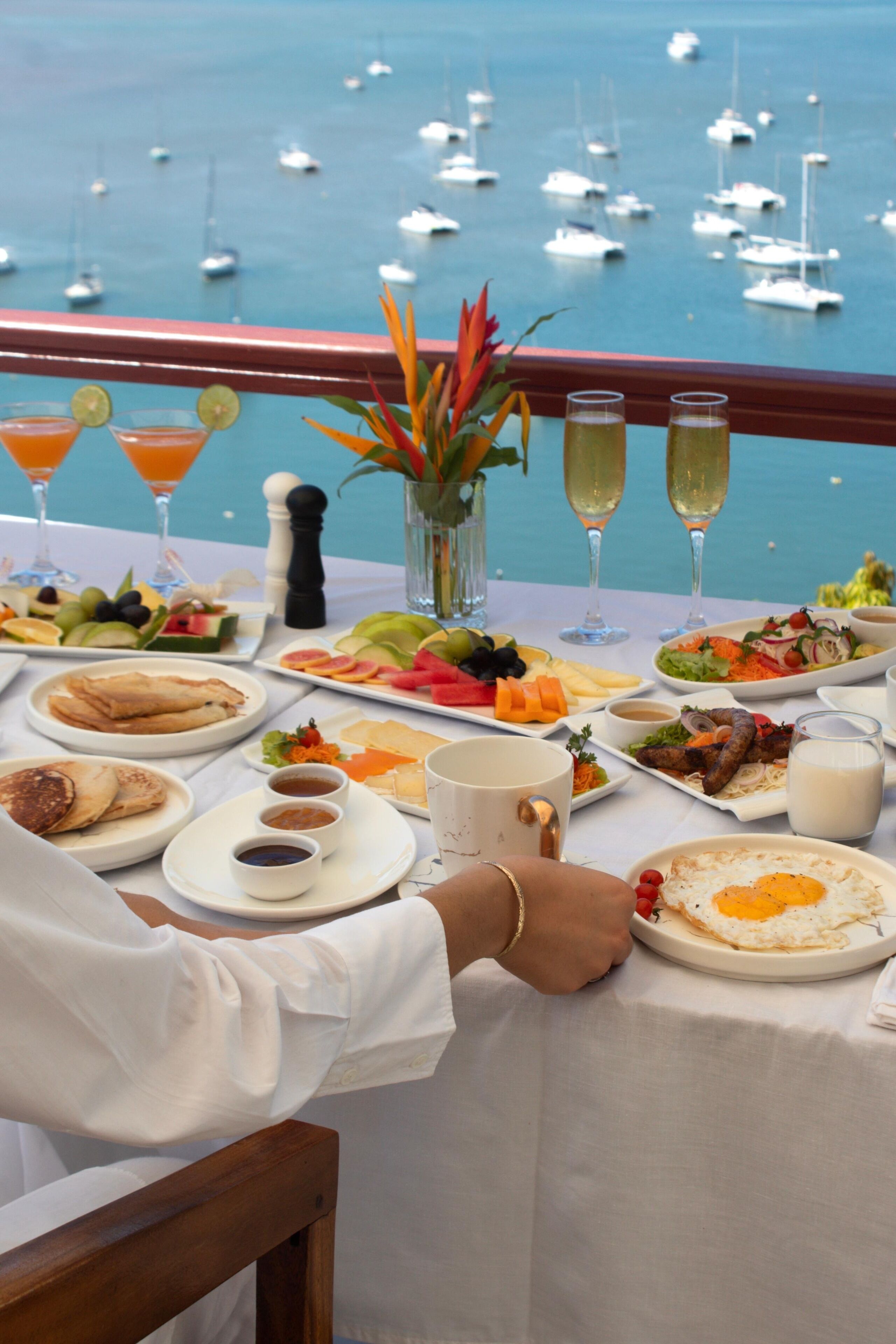 A dining table with a variety of food and drinks, overlooking a marina with boats.