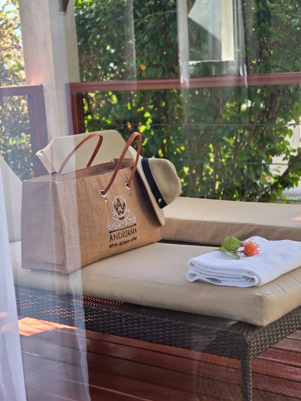 A wicker lounge chair with a beige cushion, a brown paper bag with the logo "ANDRIANA HOTEL RESORT SPA", and a folded white towel with a flower on a wooden deck.