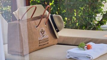 A wicker lounge chair with a beige cushion, a brown paper bag with the logo "ANDRIANA HOTEL RESORT SPA", and a folded white towel with a flower on a wooden deck.