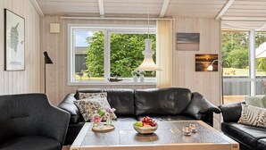 En moderne stue med en sort lædersofa, et træsofabord med pynteting og store vinduer med udsigt til grønne omgivelser.