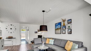 Ett modernt vardagsrum med en grå hörnsoffa, ett runt soffbord och en svart hänglampa. En vit väckarklocka är monterad på väggen, och det finns inramade tavlor ovanför soffan.