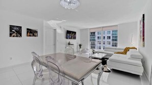Una sala de estar moderna con un sofá blanco, una mesa de comedor de vidrio y sillas transparentes. Hay un ventanal con vistas a edificios.