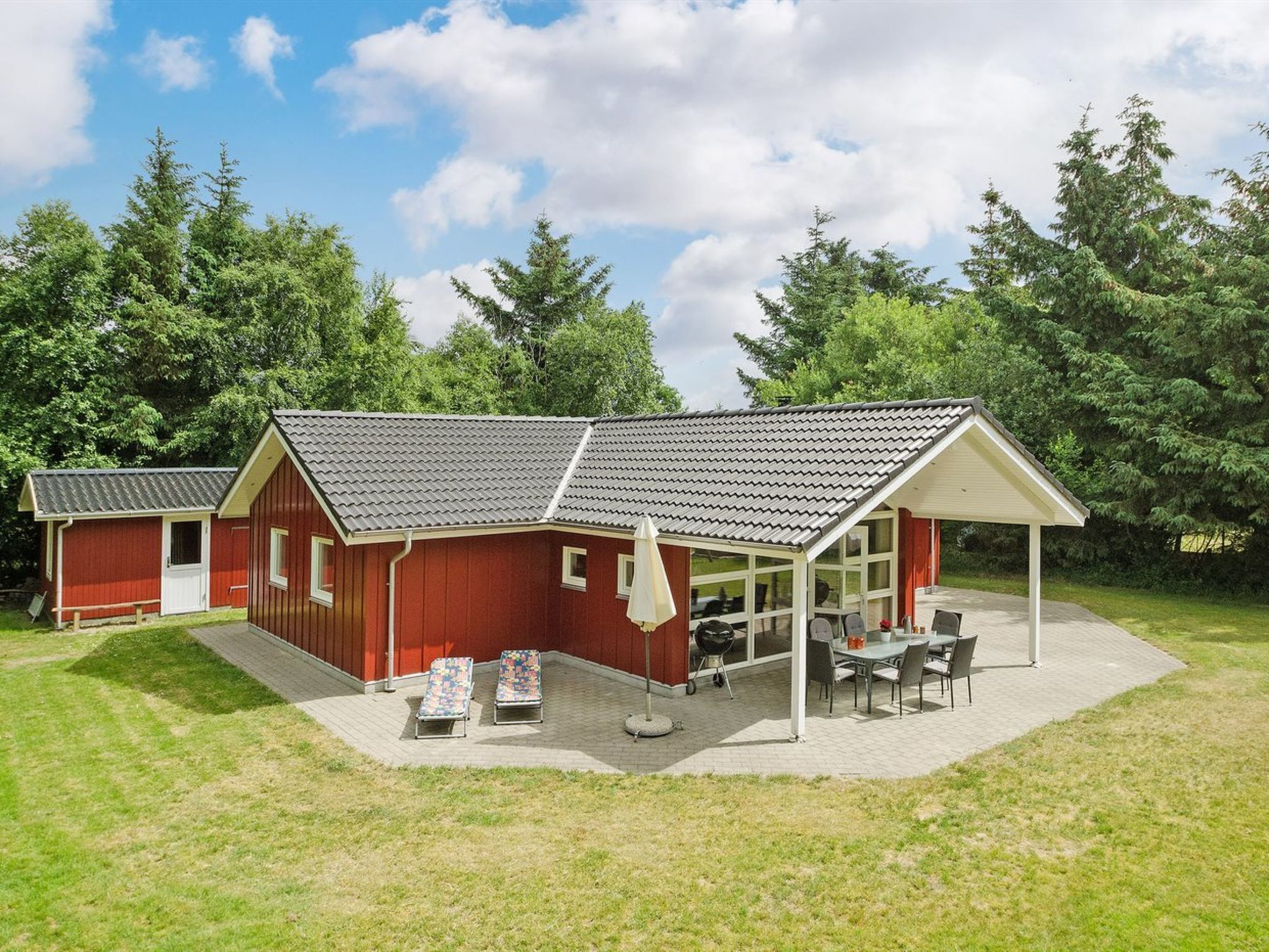 Ein rotes Gebäude mit einer überdachten Terrasse, einem Grill und zwei Liegestühlen.