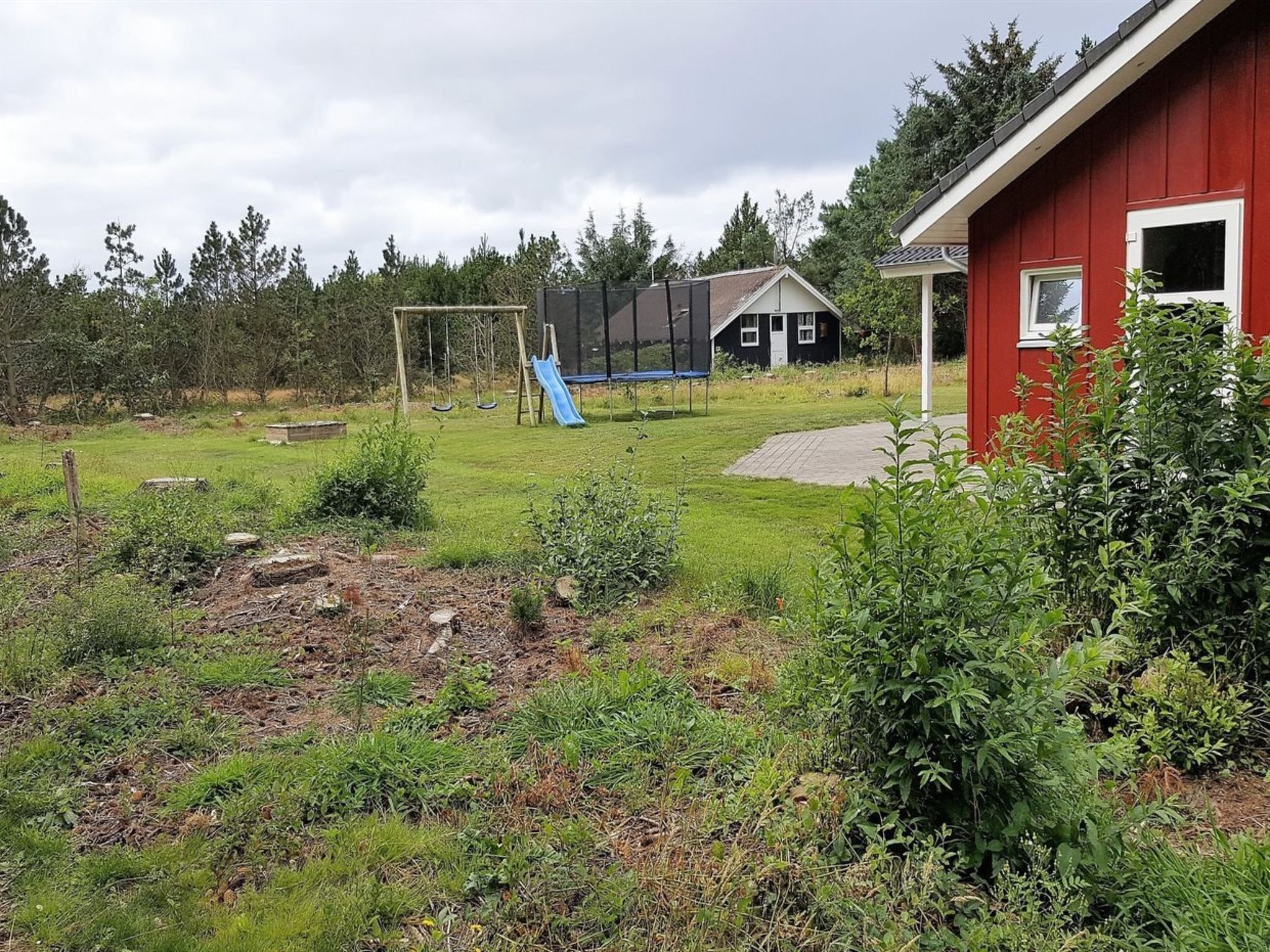 Un jardin à l’arrière d’une maison, aménagé avec un trampoline, une balançoire et un petit abri de jardin.