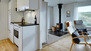 Et moderne køkken med hvide skabe, en sort bordplade og en brændeovn. Der er to træstole med hynder i et siddeområde med udsigt til det fri.