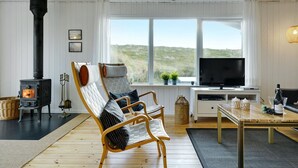 En hyggelig stue med en brændeovn, to bløde lænestole, et fjernsyn og et stort vindue med udsigt over landskabet.