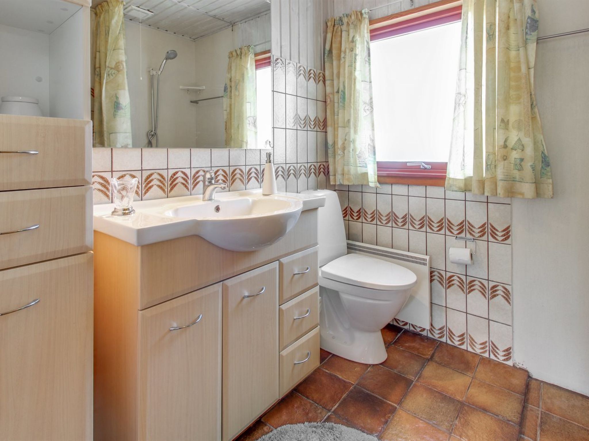 A bathroom with a sink, toilet, and shower. There are curtains on the windows and a tiled backsplash.