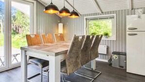 A modern kitchen with a dining area, featuring a wooden table and chairs, pendant lights, and a view of greenery outside.
