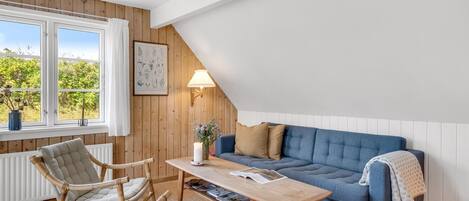 A cozy room with a blue sofa, wooden chairs, a table with books, a lamp, and a window with a view of greenery.