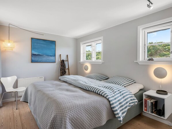 Un dormitorio con cama, mesitas de noche, una silla, un cuadro y ventanas.