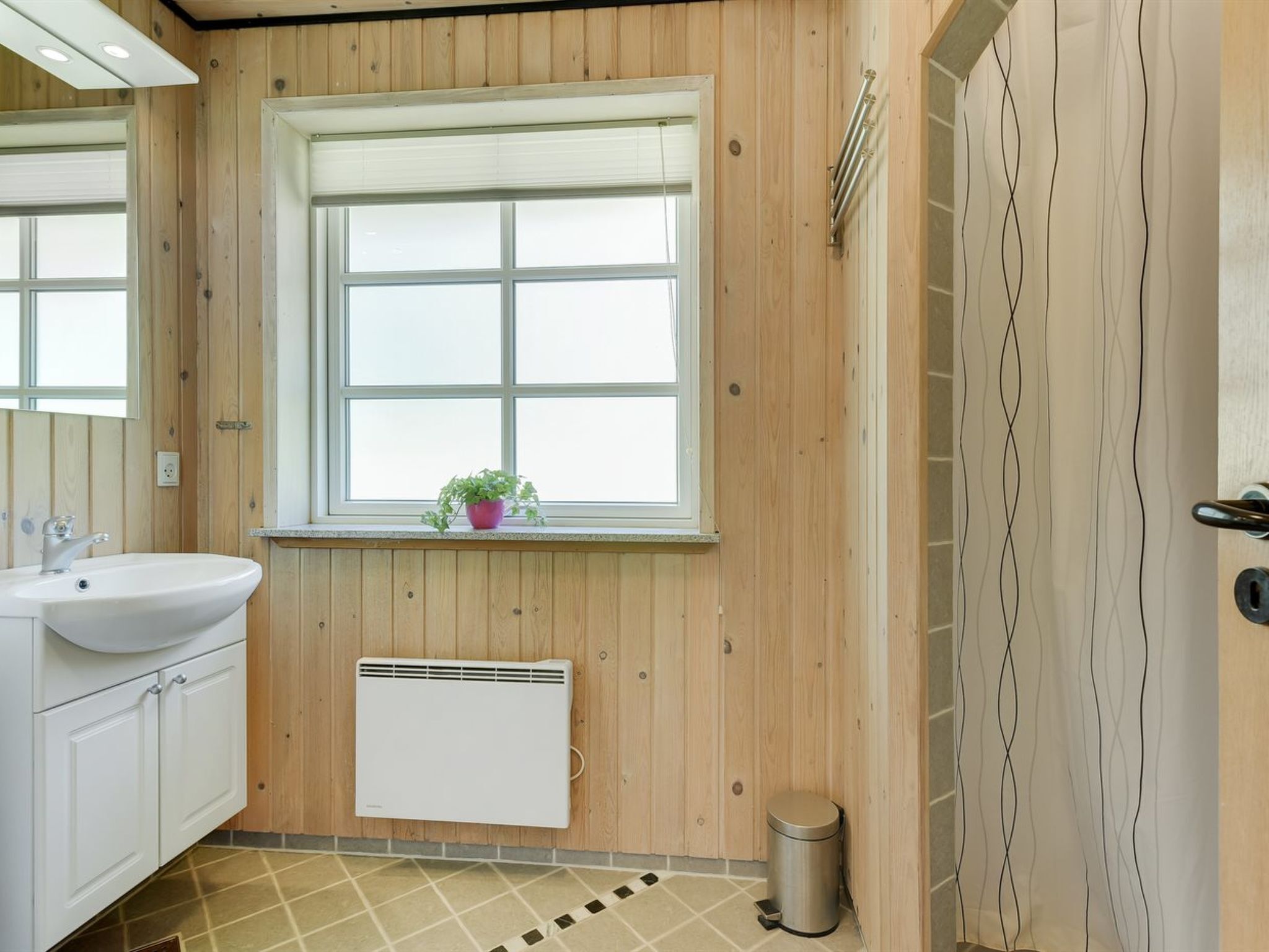 Une salle de bain avec des murs en bois, une fenêtre avec une plante, un lavabo blanc, un radiateur et une poubelle.