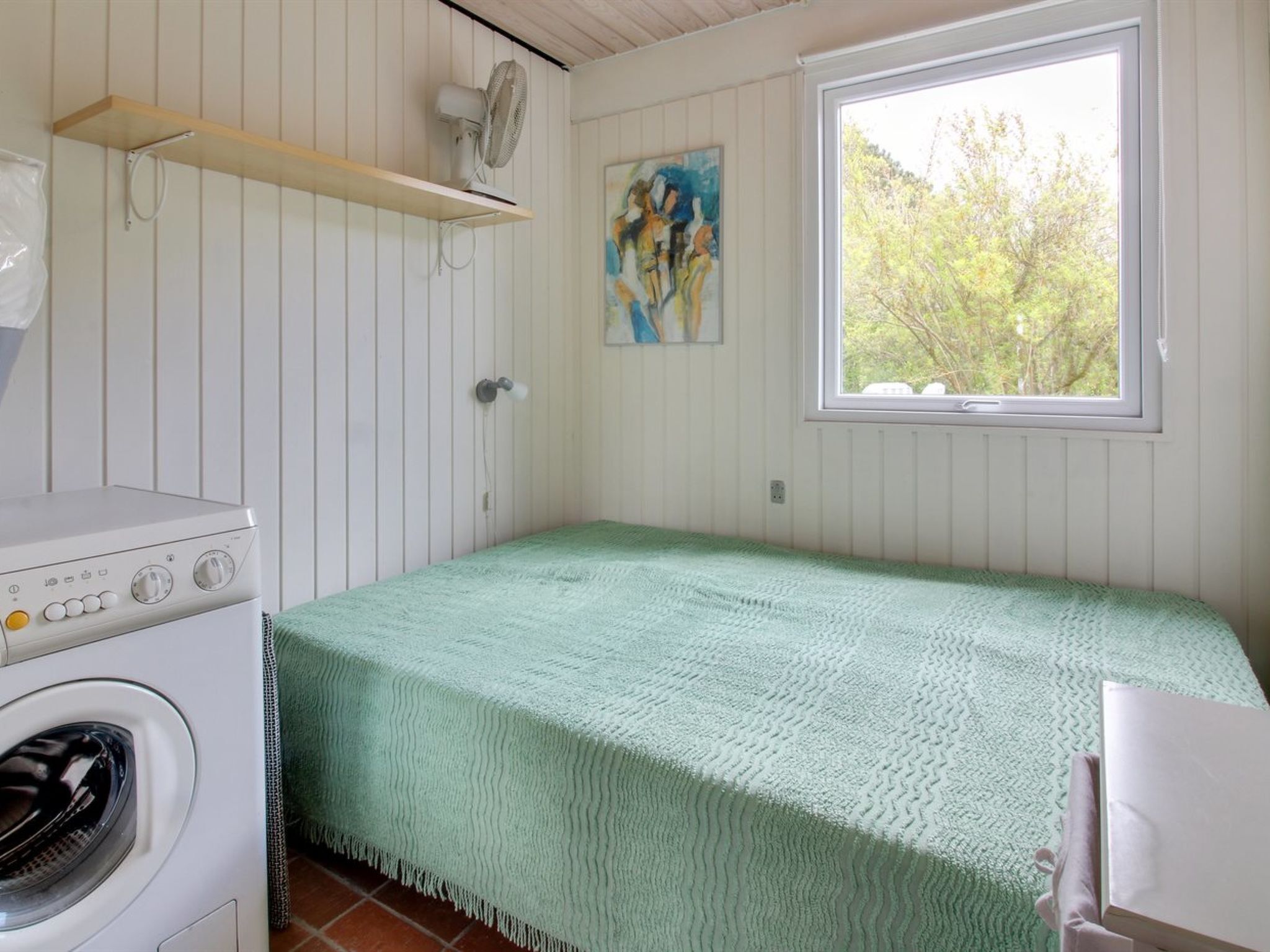 A small, single-room accommodation with a washing machine, a bed with a green blanket, a window with a view of trees, and a painting on the wall.