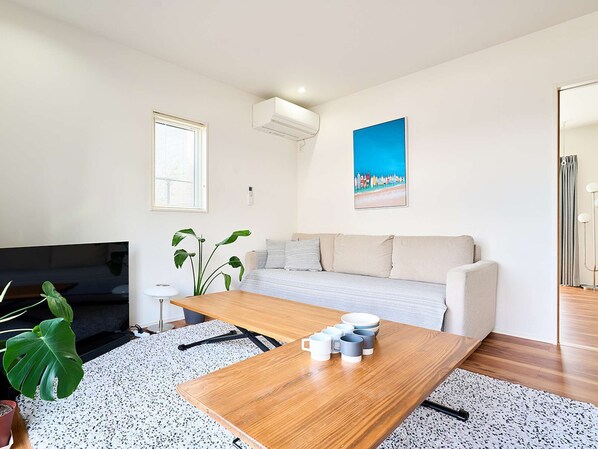 A modern living room with a sofa, a coffee table with mugs, a large plant, and a painting on the wall.