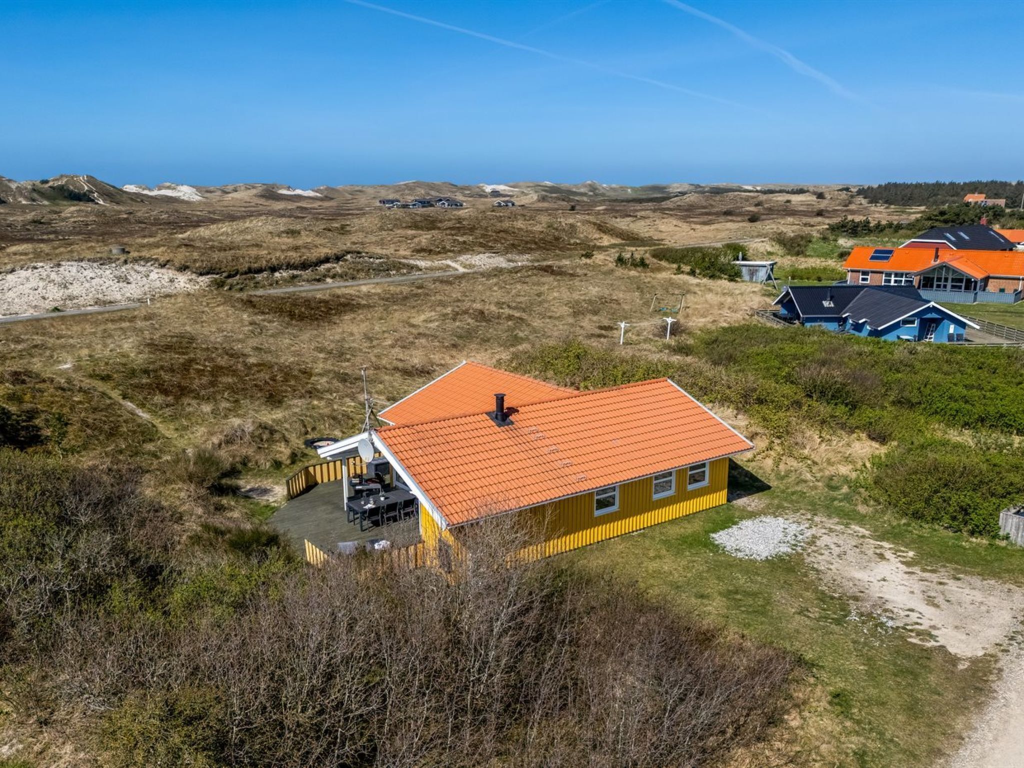 En gul bygning med rødt tag, omgivet af et naturlandskab med klitter og sparsom vegetation.