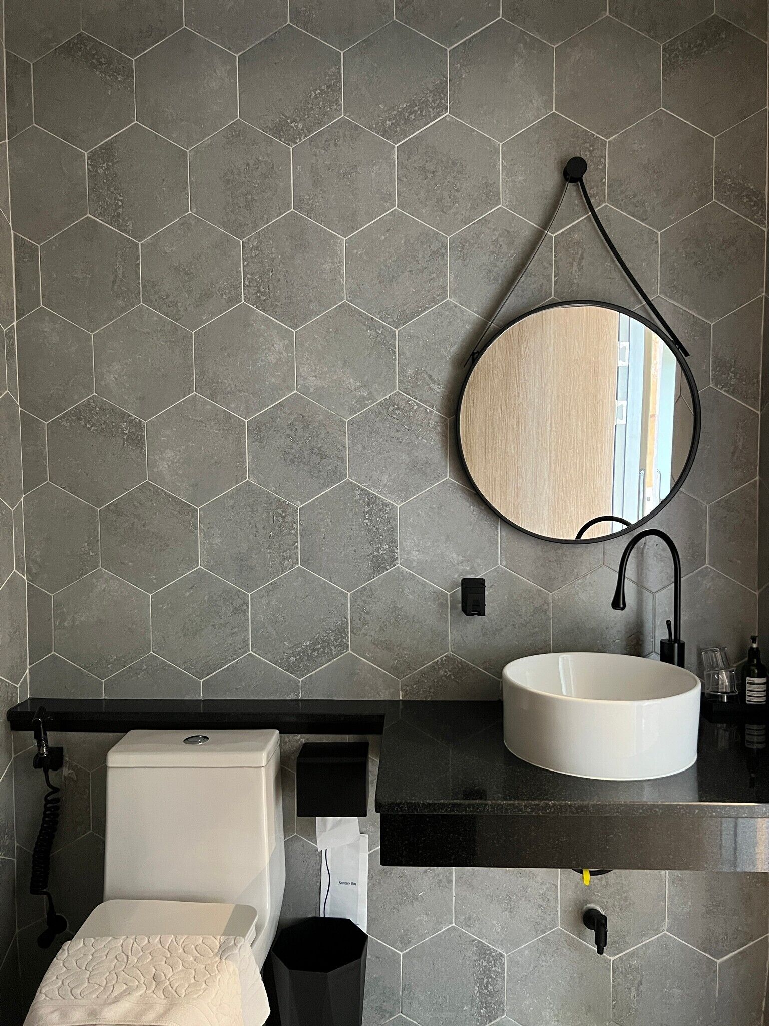 A bathroom with a hexagonal tile backsplash, a round mirror, a white sink, and a toilet.