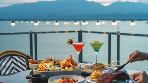 Una mesa servida con diversos platos y bebidas, con vistas a un cuerpo de agua y montañas al fondo.