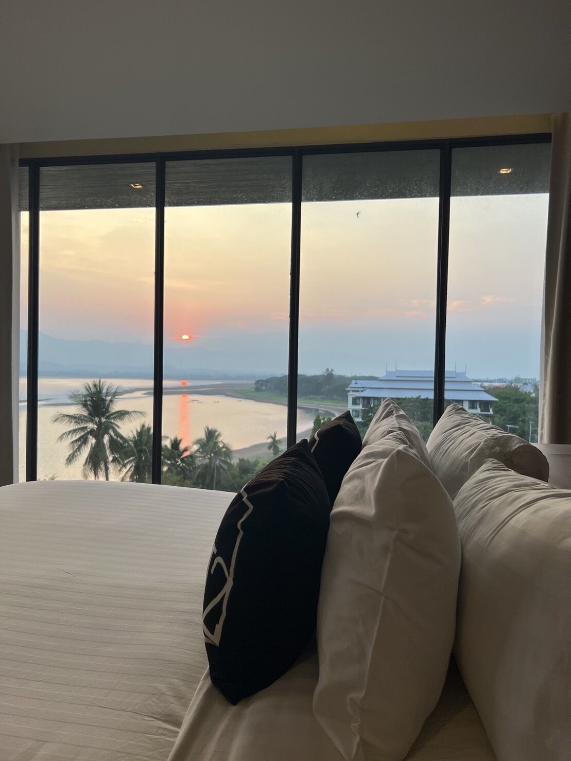 Ein Schlafzimmer mit großem Fenster, das Blick auf einen Sonnenuntergang, einen Strand und ein Gebäude bietet.