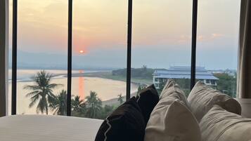 Ein Schlafzimmer mit großem Fenster, das Blick auf einen Sonnenuntergang, einen Strand und ein Gebäude bietet.