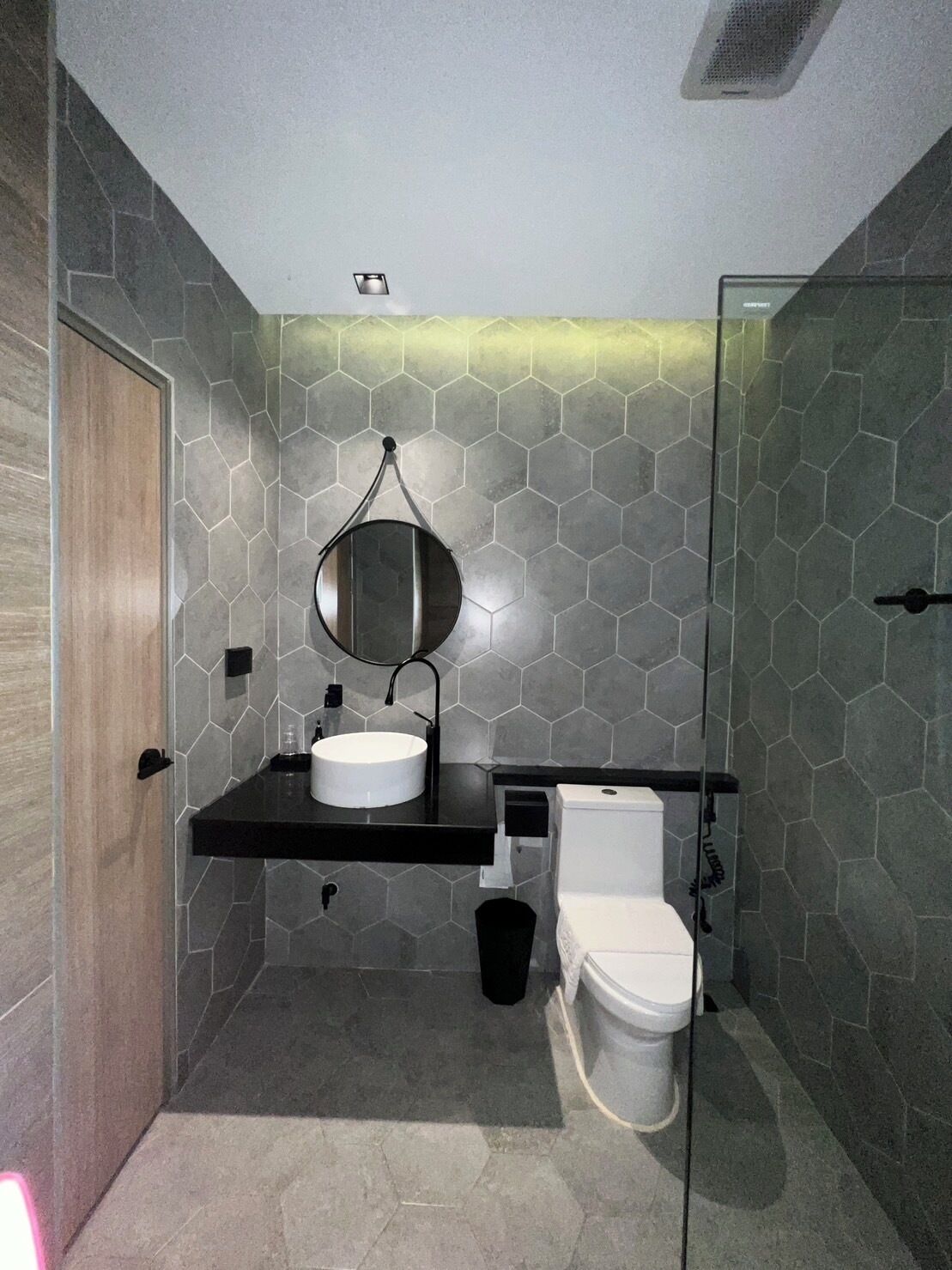 Un baño moderno con pared de azulejos hexagonales, un espejo redondo, un lavabo blanco y un inodoro.