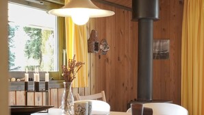 A wooden interior with a dining table, chairs, a vase with dried flowers, candles, and a large stove.