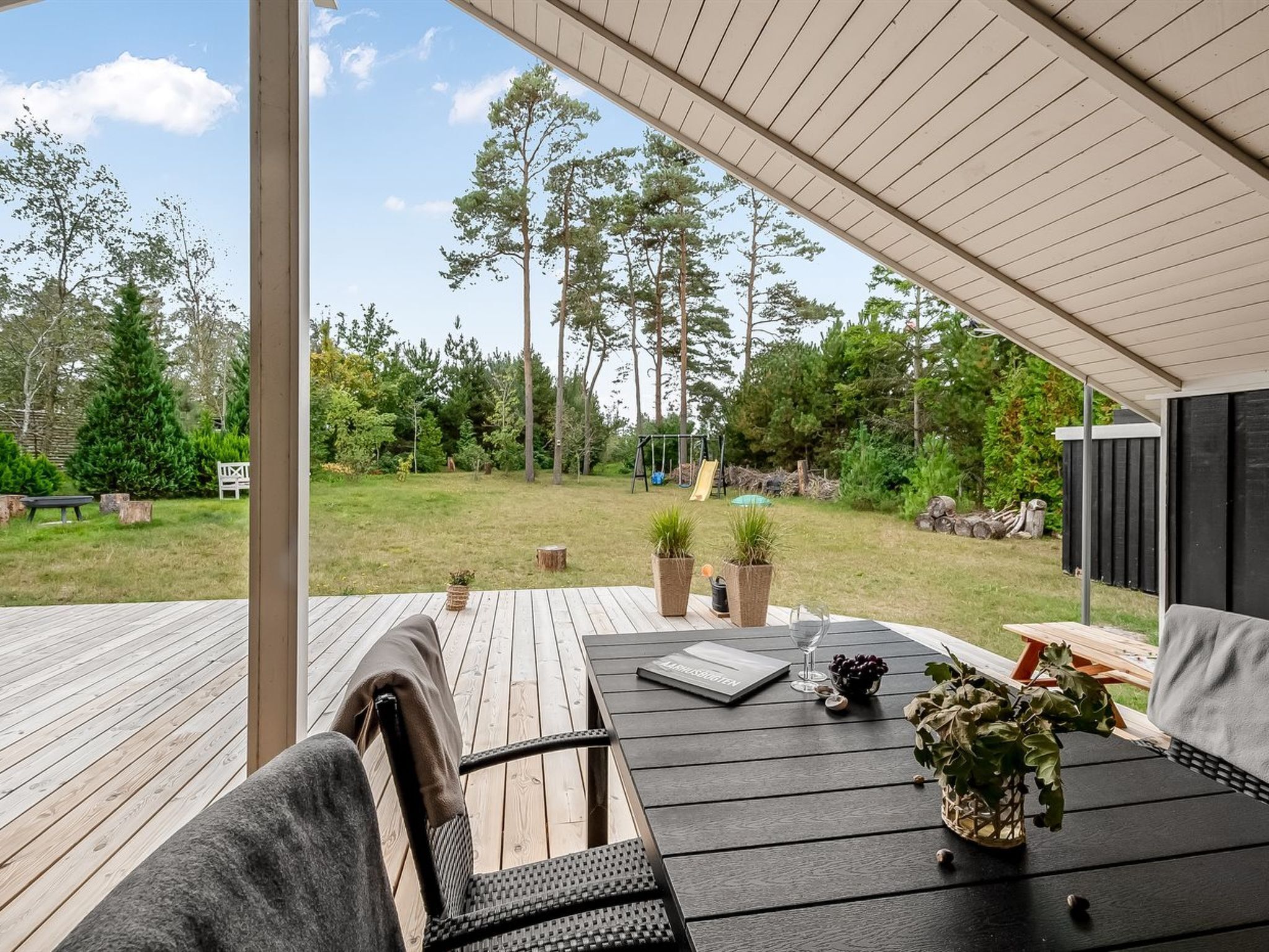 A covered outdoor seating area with a wooden deck, a table with a book and a glass, and a chair with a cushion.