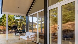 Un espace extérieur abrité doté d’une terrasse en bois, d’un fauteuil en osier et d’une porte vitrée donnant sur une vue sur les arbres et un bâtiment.