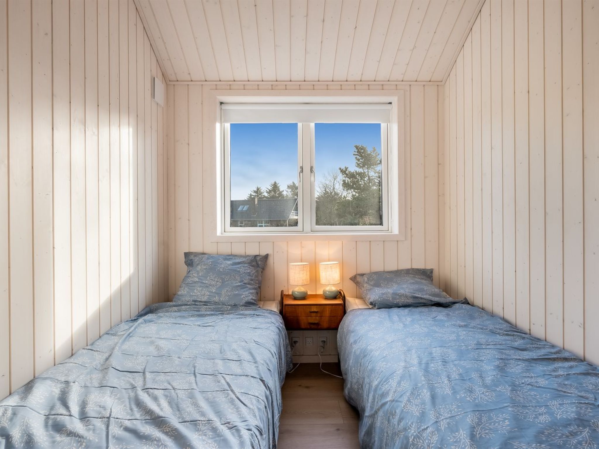 Un dormitorio pequeño con dos camas individuales, una mesita de noche con lámparas y una ventana con vistas a árboles.