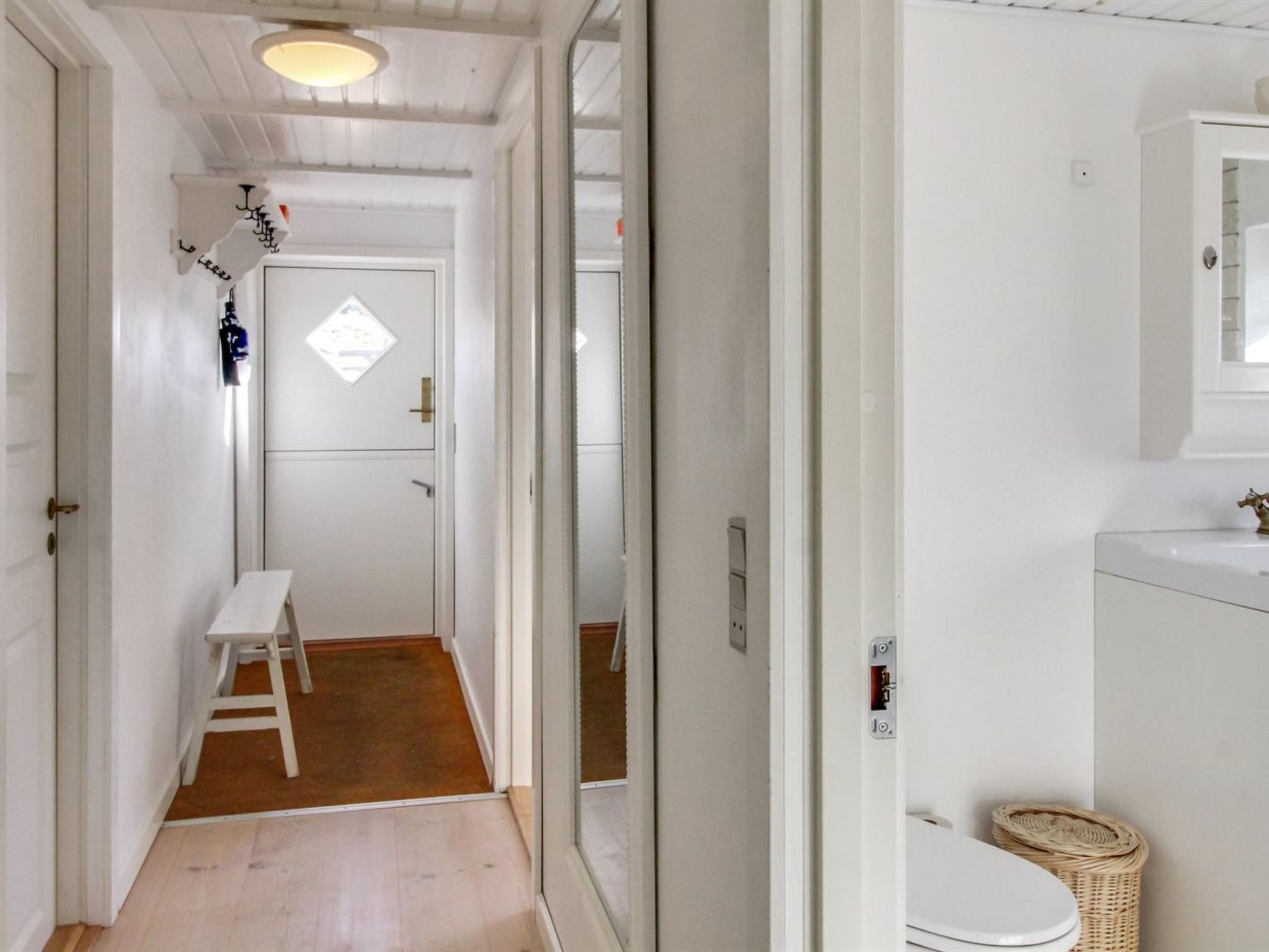 A narrow hallway with a wooden floor leading to a white door with a small window. A white stool is placed against the wall, and a wicker basket is on the floor.