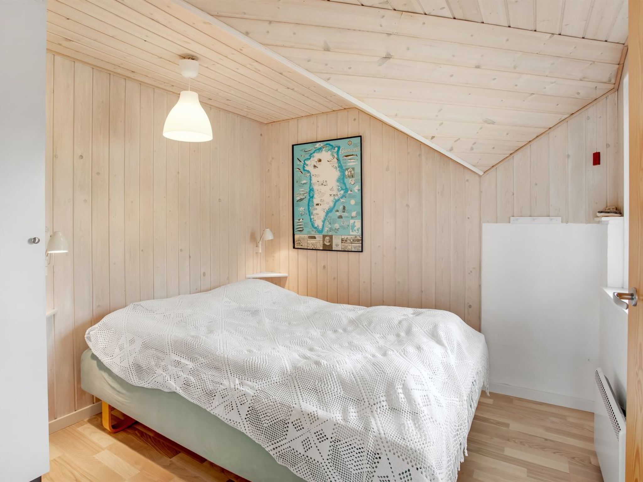 Une chambre avec un plafond en bois, un lit recouvert d’une couverture blanche tricotée, une carte accrochée au mur et un radiateur blanc.