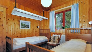 A wooden cabin room with two single beds, a chair, a heater, a window with curtains, and a framed picture on the wall.
