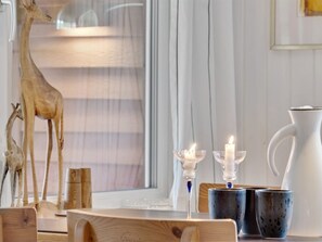 Een kamer met een houten tafel, een witte karaf, twee kaarsen in glazen houders en een groot raam met dunne gordijnen.