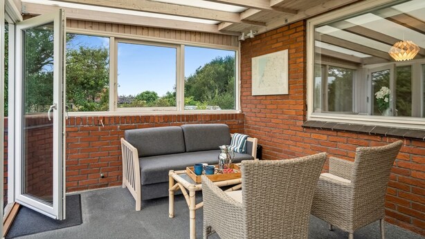 A cozy outdoor seating area with a sofa and wicker chairs, a table with a tray of items, and a view of greenery through large windows.