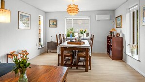 A dining area with a wooden table, chairs, and a chandelier. There are framed pictures on the walls and a window with blinds.