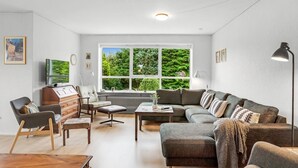 A modern living room with a sofa, chairs, a coffee table, and a TV. There is a large window with a view of trees.