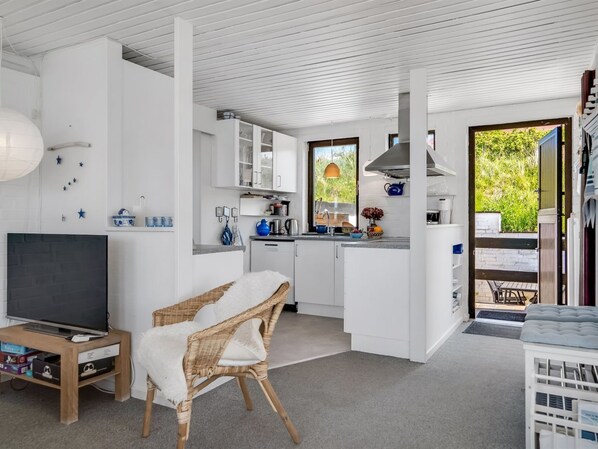 A modern kitchen with white cabinetry, a dining area with a wicker chair, and a television on a wooden stand.