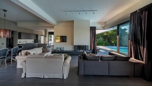 A modern living room with a large grey sofa, a dining area with a kitchen in the background, and a view of a swimming pool through large windows. - Minimal brand new villa with private pool close to Athens (Kalamos)