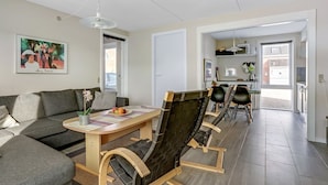 A modern living room with a sofa, a dining table, and chairs. There is a painting on the wall, a ceiling fan, and a view into the kitchen.