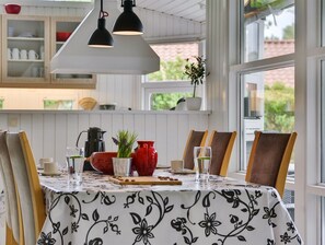 A dining area with a table set for four, featuring a floral tablecloth, wooden chairs, and a view of the outdoors through large windows.