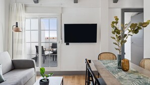 Ein modernes Wohnzimmer mit Sofa, Esstisch und Stühlen, einem flachbildfernseher an der Wand und einem großen Fenster mit Blick ins Freie.