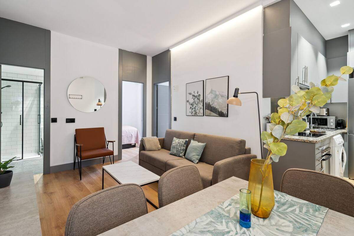 Ein modernes Wohnzimmer mit Sofa, Couchtisch und Essbereich. Eine Küche mit sichtbarer Mikrowelle und Spüle ist ebenfalls vorhanden.