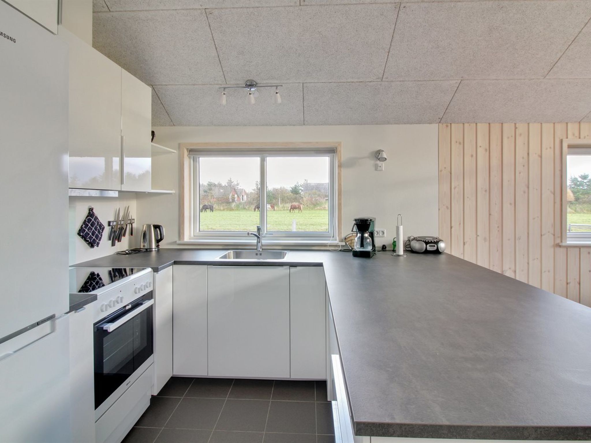 A modern kitchen with white cabinets, a built-in oven, a large window, and a view of a green landscape.