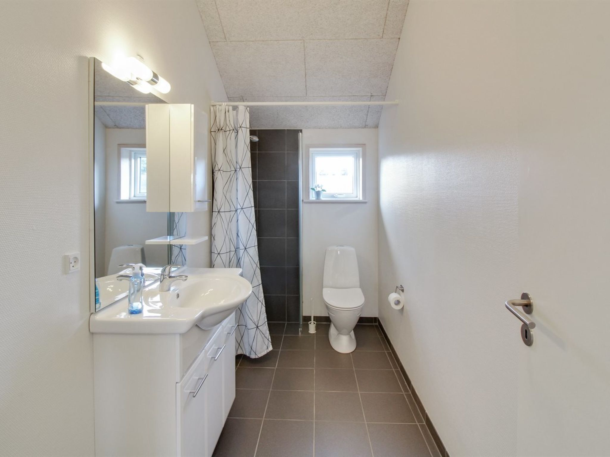 A compact bathroom with a white sink, mirror, and toilet. A shower curtain with a geometric pattern is visible, and a window above the toilet allows natural light.