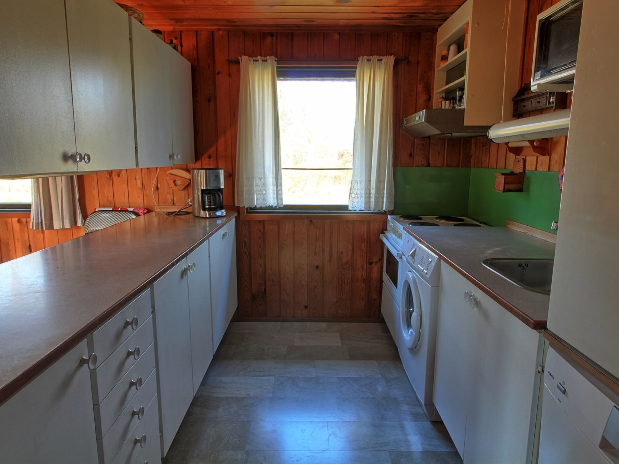 Ett kompakt kök med träväggar, en tvättmaskin, en kaffebryggare och ett fönster med gardiner.